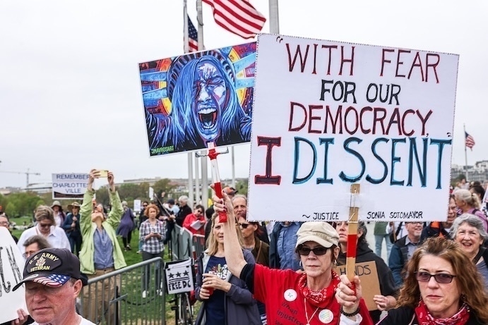 A group of people at a demonstration hold signs, one with a painted face and another reading WITH FEAR FOR OUR DEMOCRACY I DISSENT.
