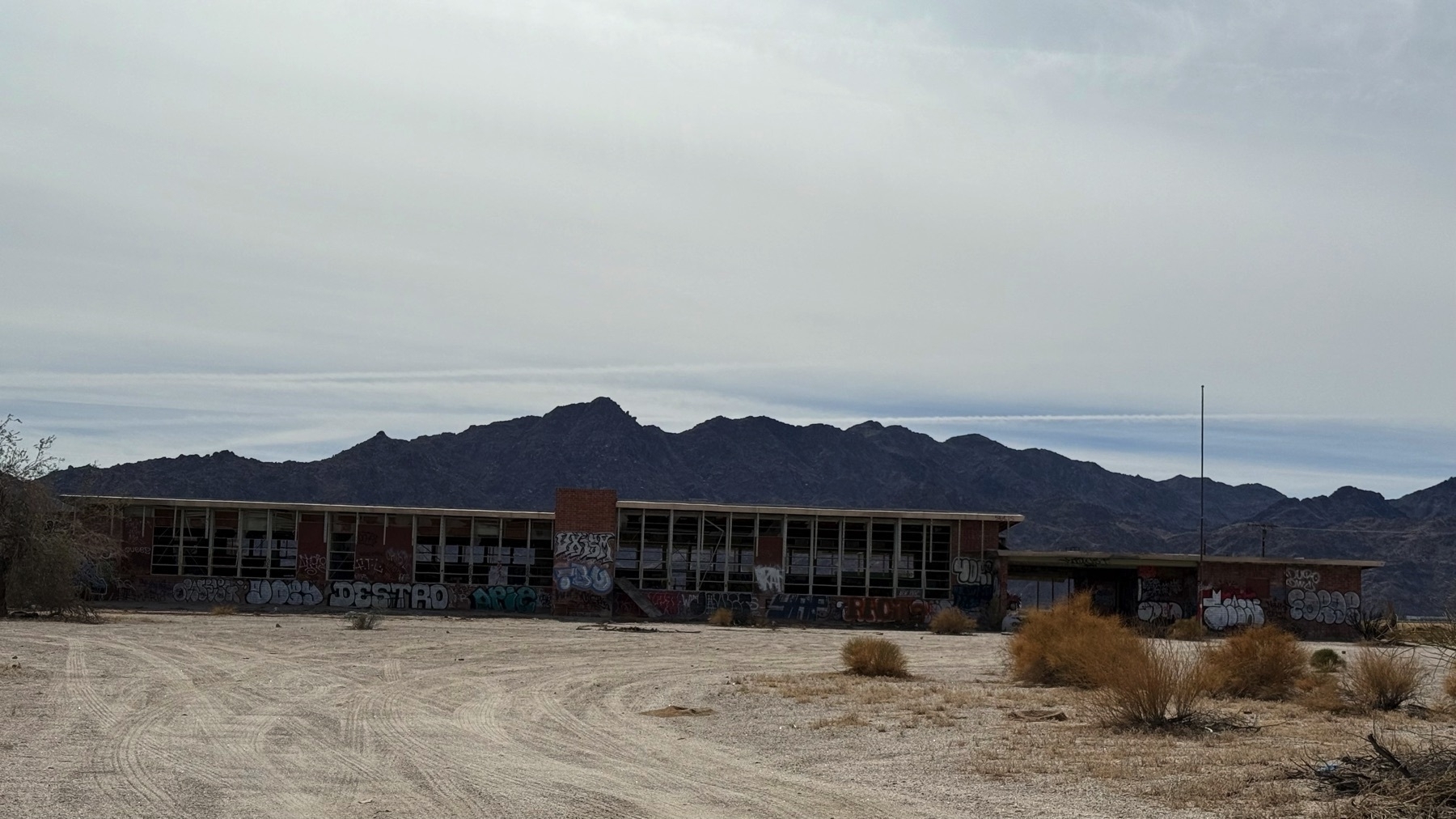 A long, abandoned building covered in graffiti stands in a barren, desert landscape with distant mountains in the background.