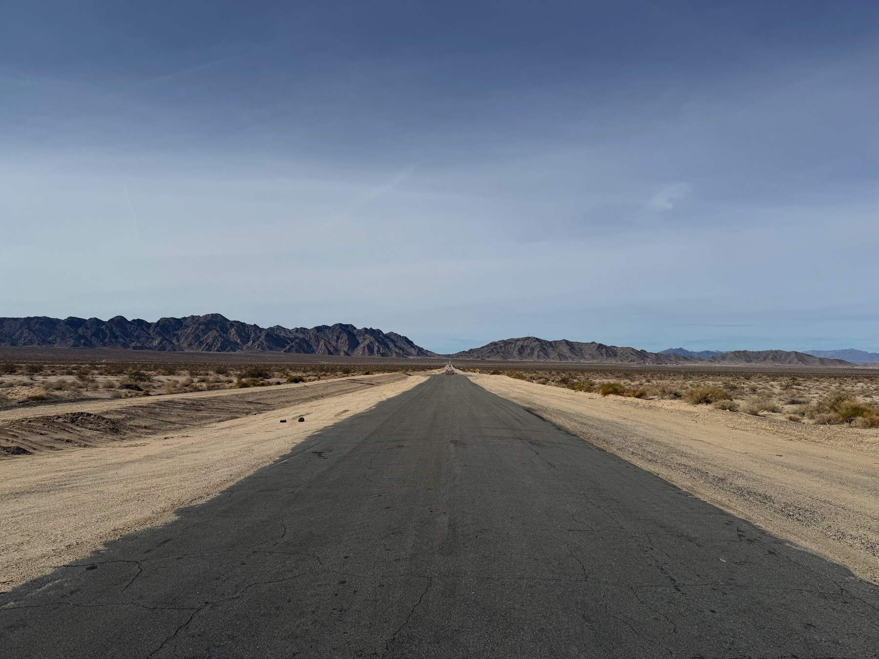 A long, straight road stretches through a barren desert landscape with mountains in the distance.