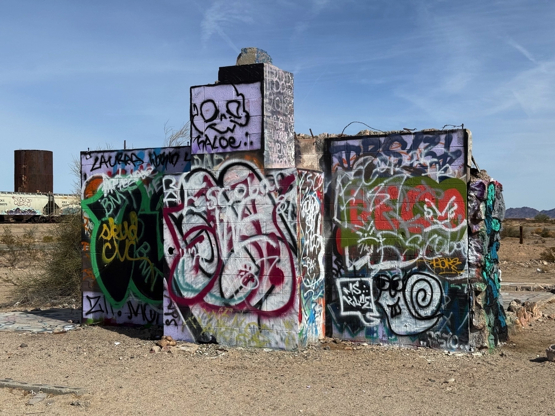 A heavily graffiti-covered concrete structure stands in a barren outdoor setting under a blue sky.